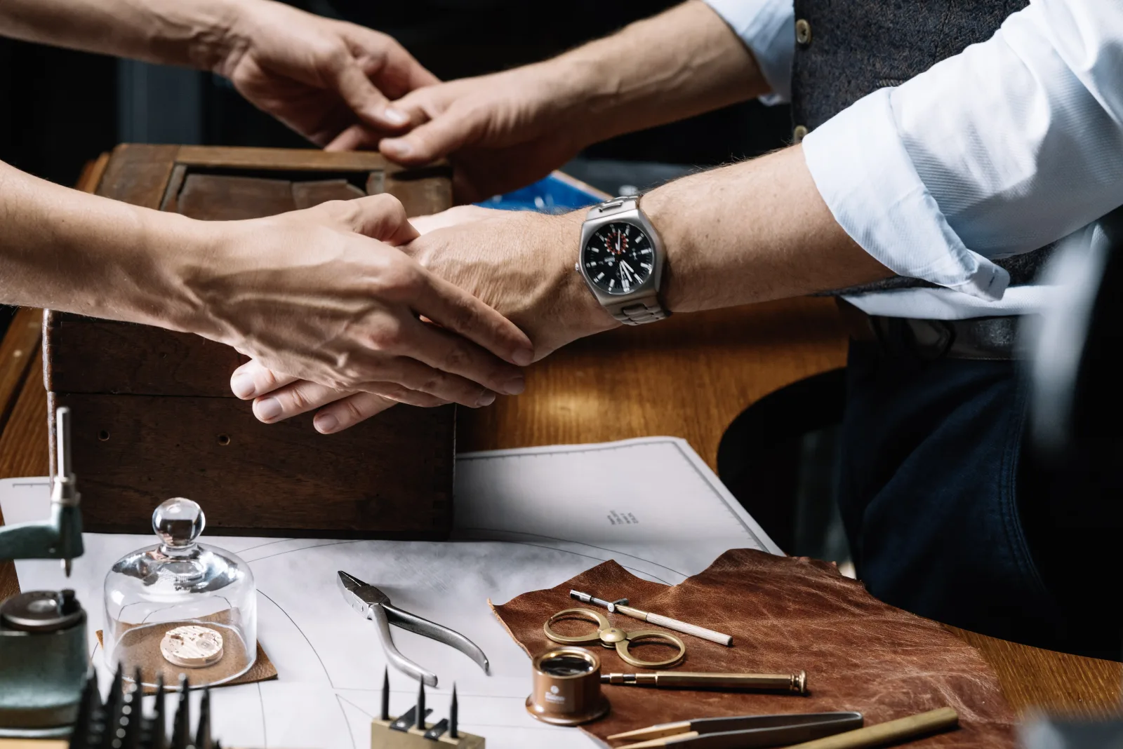 Hands at watchmaker desk with bell jar, compass, and precision tools — what coming home built